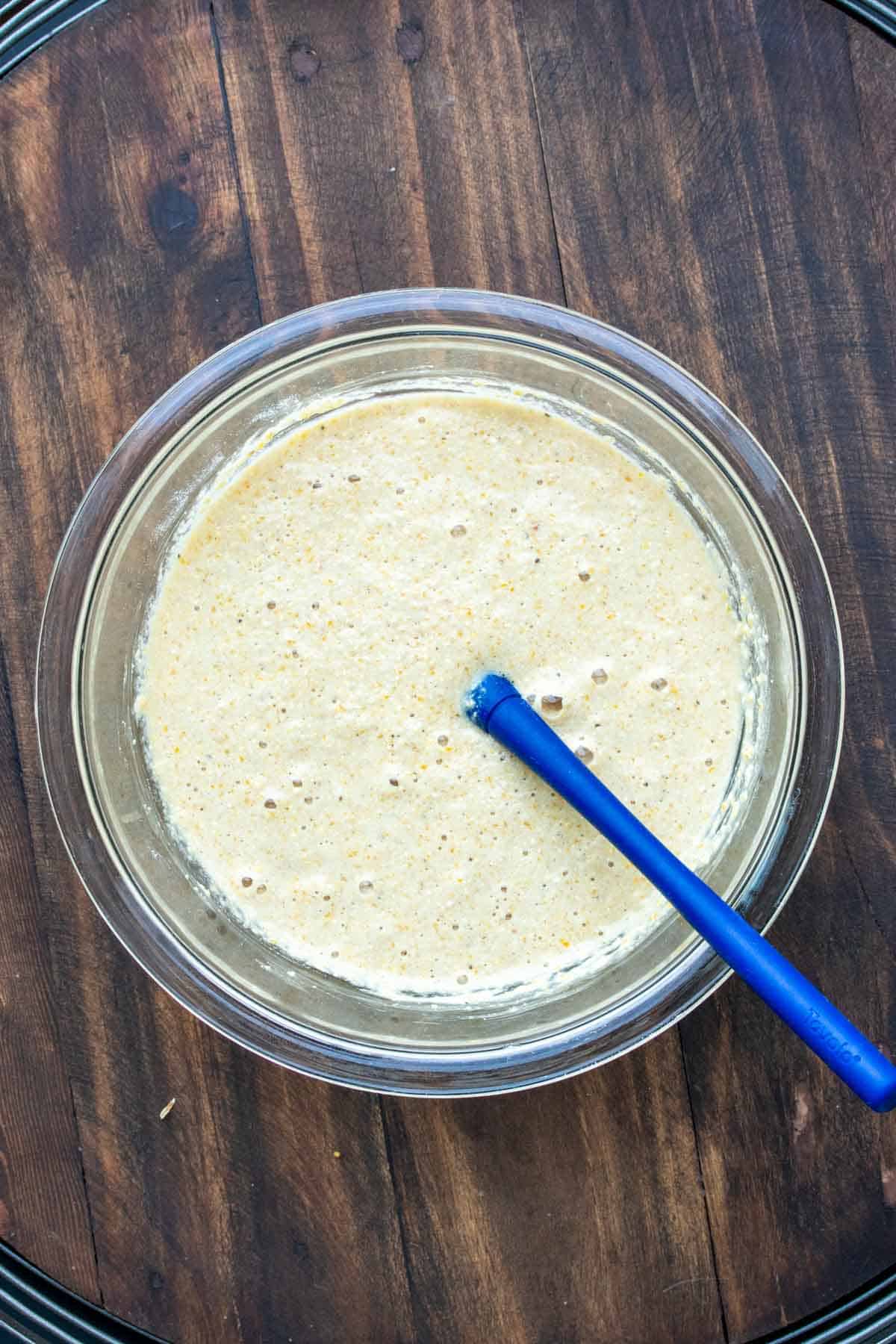 Blue spatula mixing cornbread batter in a glass bowl