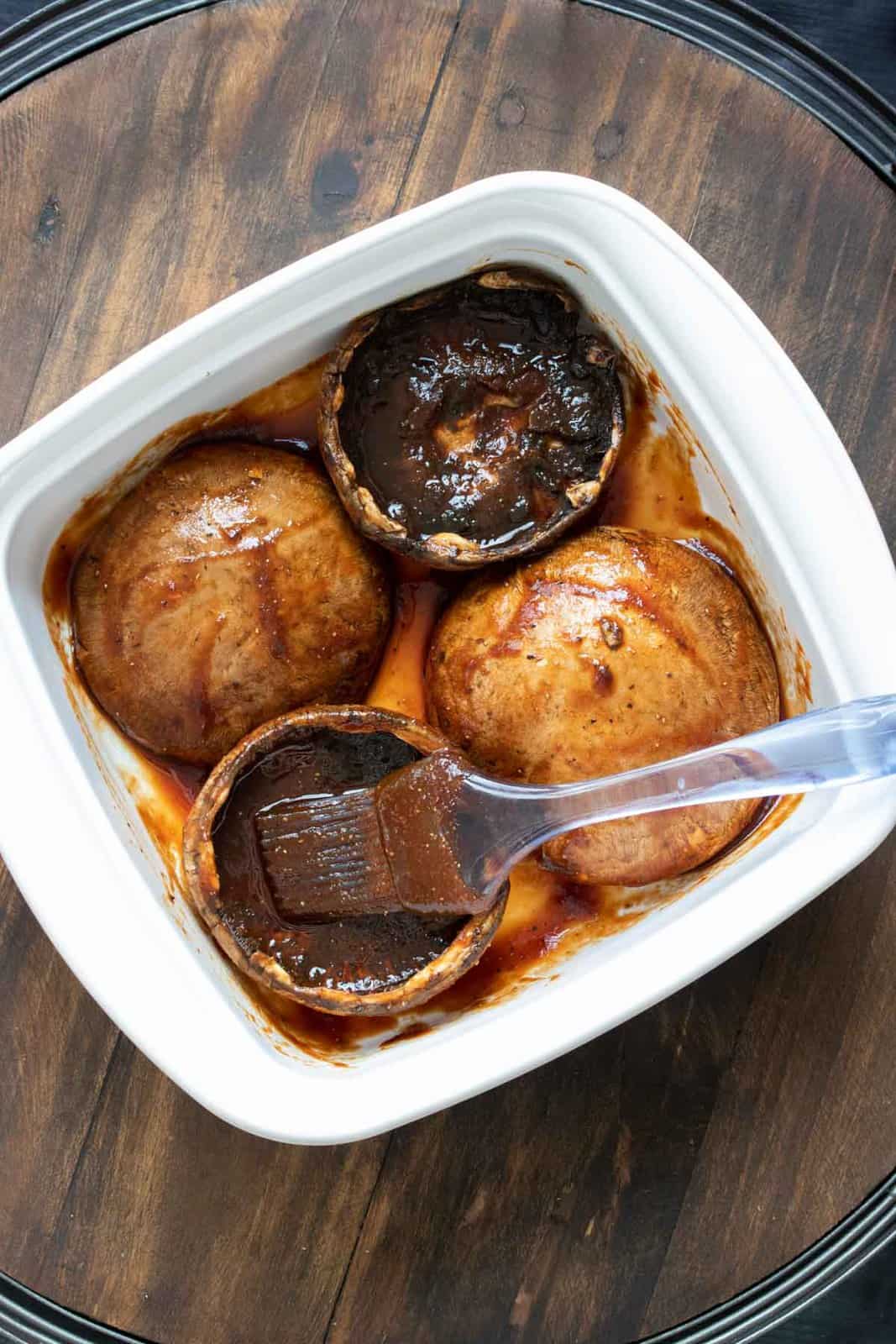 Four portobello mushrooms in a white baking dish being brushed with red sauce
