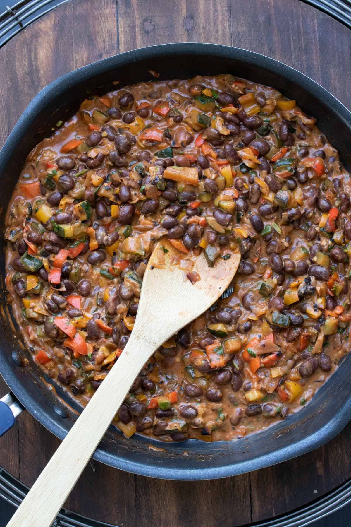 Wooden spoon mixing black bean mixture with veggies in a pan.