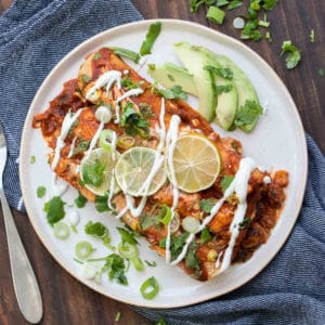 A white plate with two enchiladas on it topped with sour cream, limes and green onions.