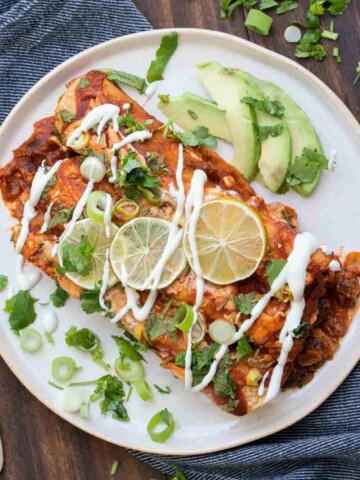 A white plate with two enchiladas on it topped with sour cream, limes and green onions.