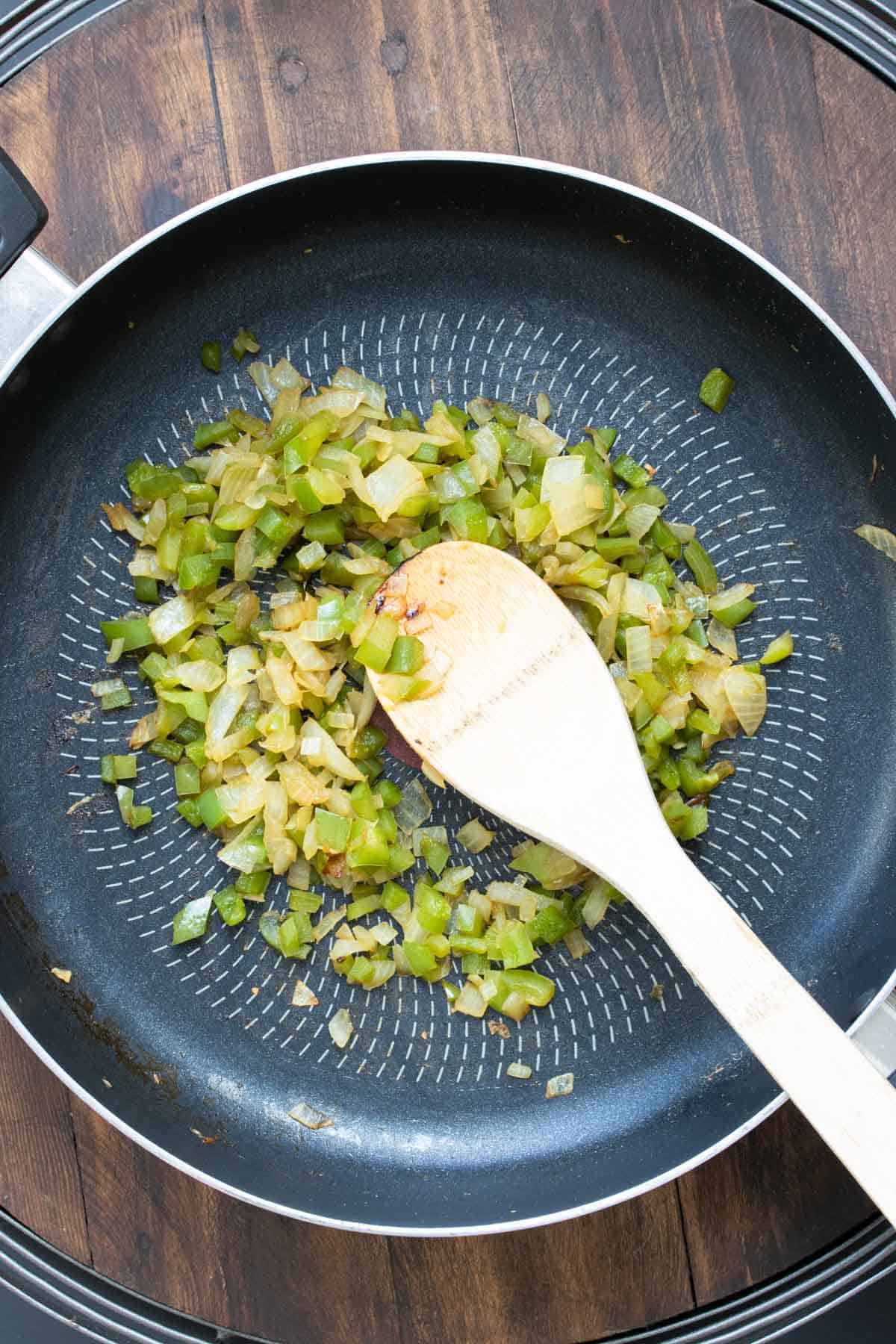 Wooden spoon sautéing onions and peppers in a pan