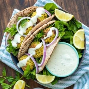 A black wide bowl with two pita falafel sandwiches in it with parsley sprigs, lemon slices and tzatziki dip.