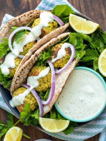 A black wide bowl with two pita falafel sandwiches in it with parsley sprigs, lemon slices and tzatziki dip.