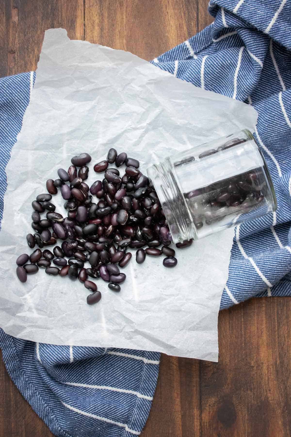 Cooked black beans spilling out of a glass jar onto parchment paper on a blue striped towel.