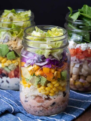 Three jars filled with salad ingredients sitting on a blue striped towel