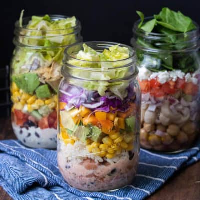 Three jars filled with salad ingredients sitting on a blue striped towel