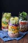 Three jars with different layered salads inside them sitting on a blue striped towel.