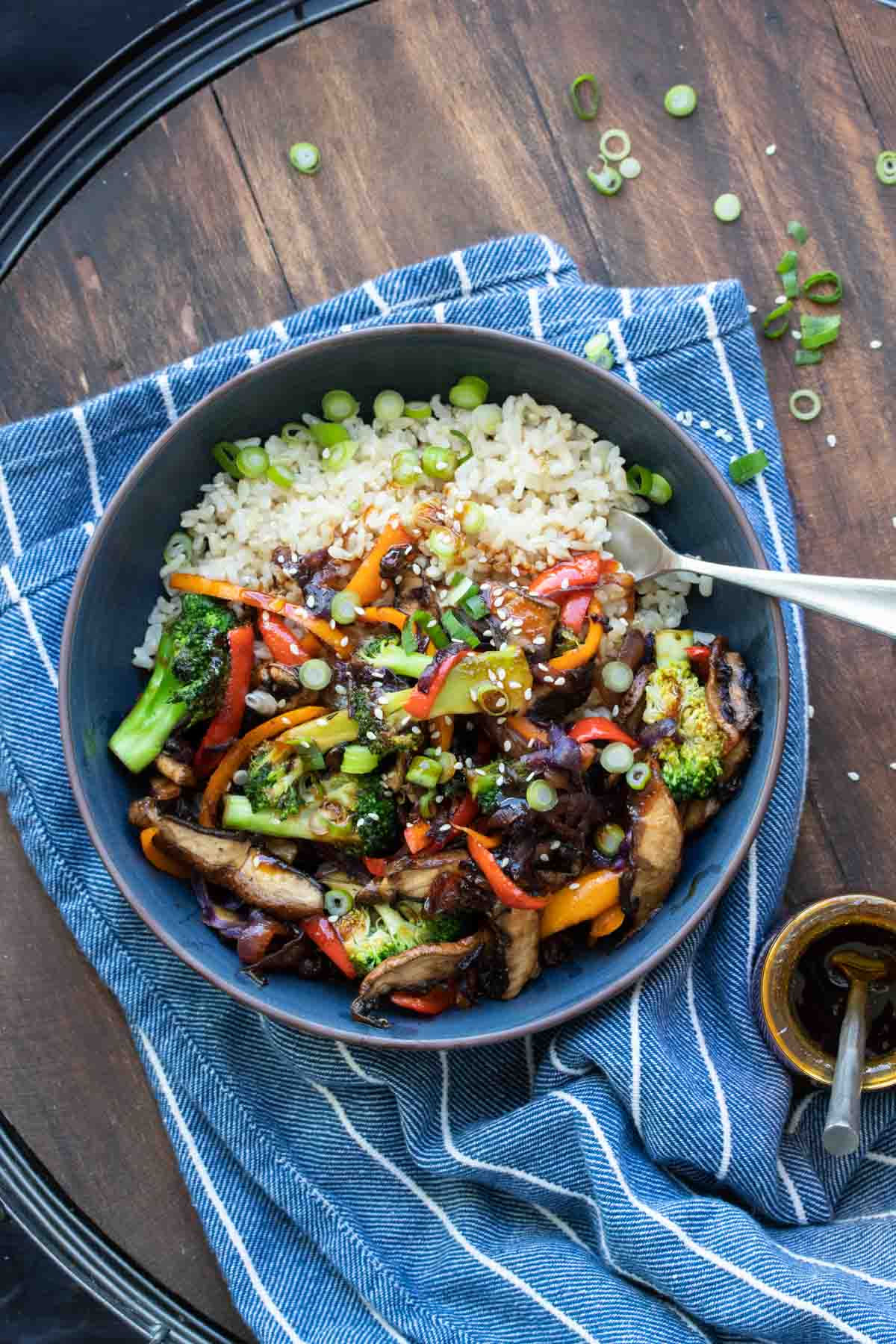 Black bowl filled with rice topped with colorful teriyaki vegetables and sliced green onions.