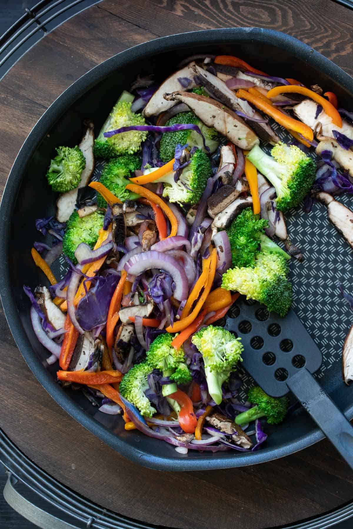 Spatula mixing colorful sliced veggies in a black pan.
