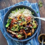 A wooden surface with a blue and white striped towel with a bowl on it filled with teriyaki vegetables over rice.