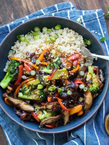 A wooden surface with a blue and white striped towel with a bowl on it filled with teriyaki vegetables over rice.