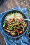 A bowl of teriyaki vegetables over rice with sliced green onions on top sitting on a blue towel on a wooden surface.