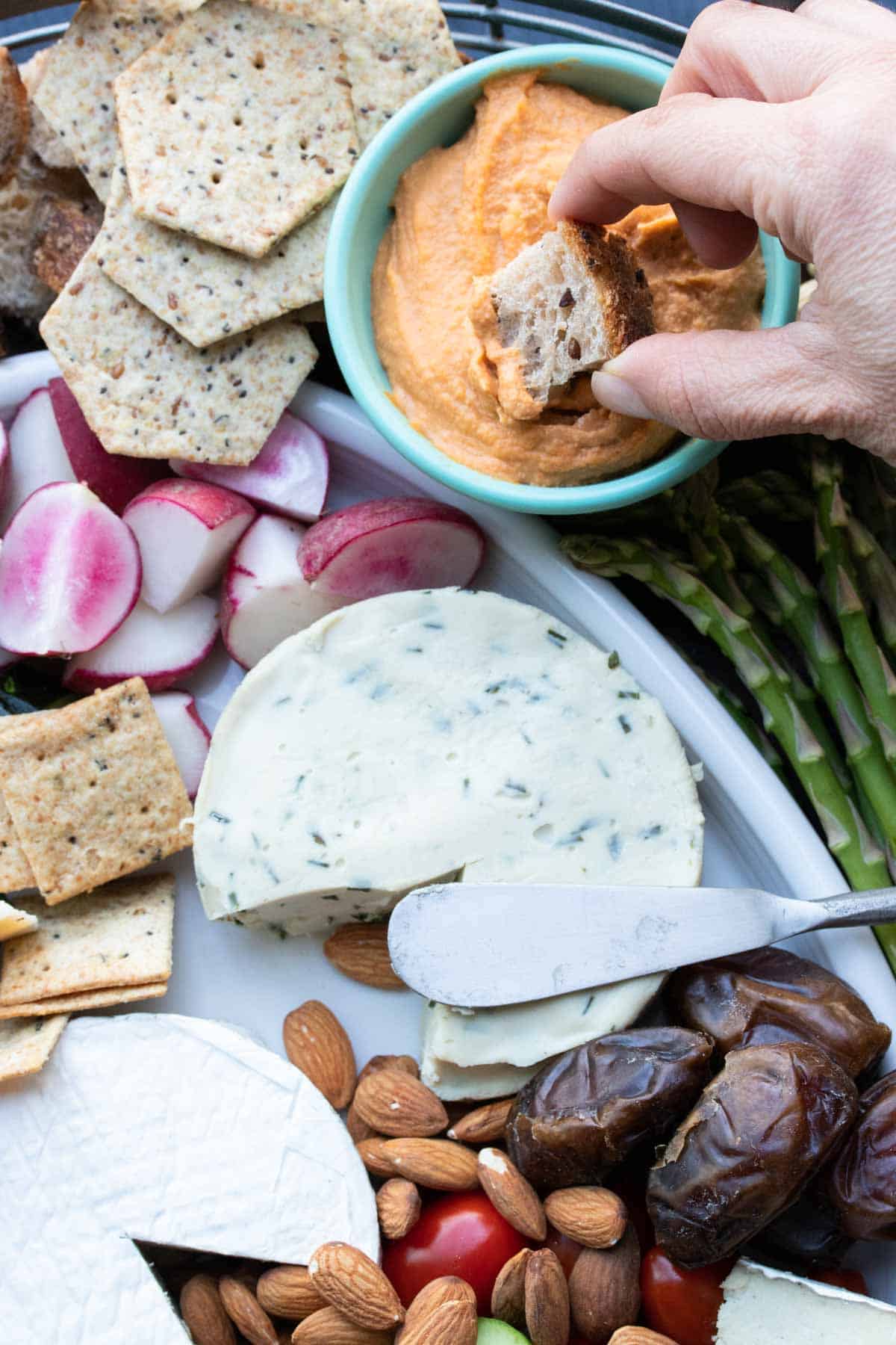 Hand dipping a piece of bread into a red hummus dip next to an appetizer platter.