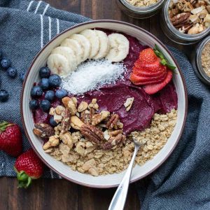 A purple smoothie bowl in a cream bowl topped with fruit, granola and nuts on a dark grey blue towel.