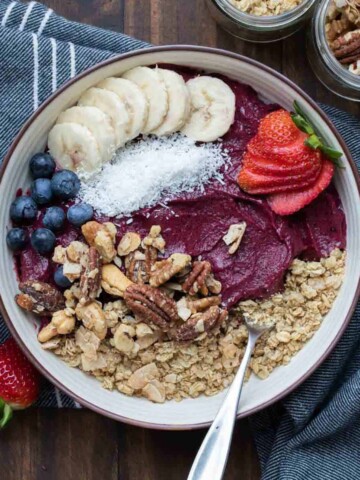 A purple smoothie bowl in a cream bowl topped with fruit, granola and nuts on a dark grey blue towel.