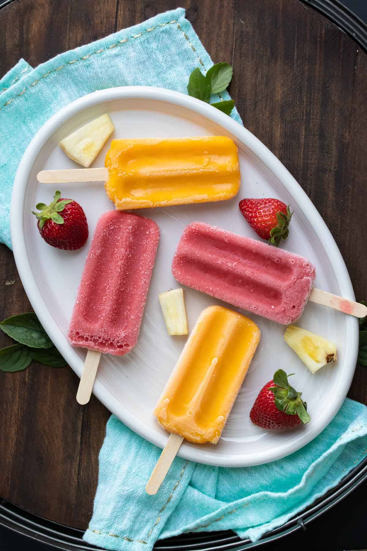 Pink and orange popsicles laying on a white plate with fruit