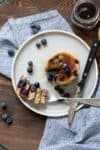 Blueberry pancakes on a cream plate with a stack of bites out on a fork laying on the plate with a knife.