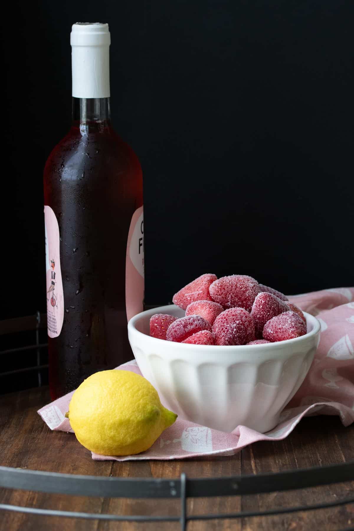 White bowl with frozen strawberries next to a lemon and wine bottle.