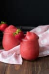 Frozen rose drinks in glass jars with strawberries on the rim near a pink towel.