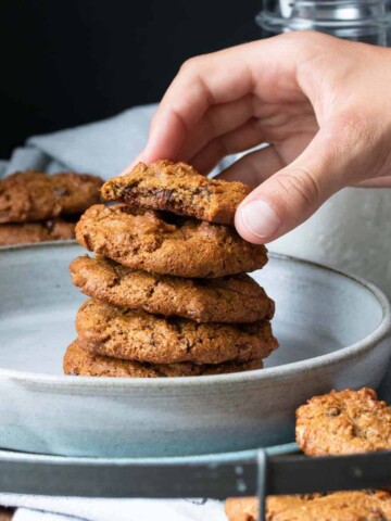 A hand taking the top chocolate chip cookie from a stack of five on a plate