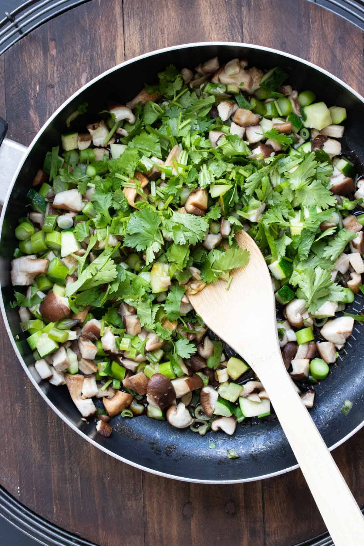 Chopped mushrooms, asparagus and cilantro being mixed in a pan by a wooden spoon