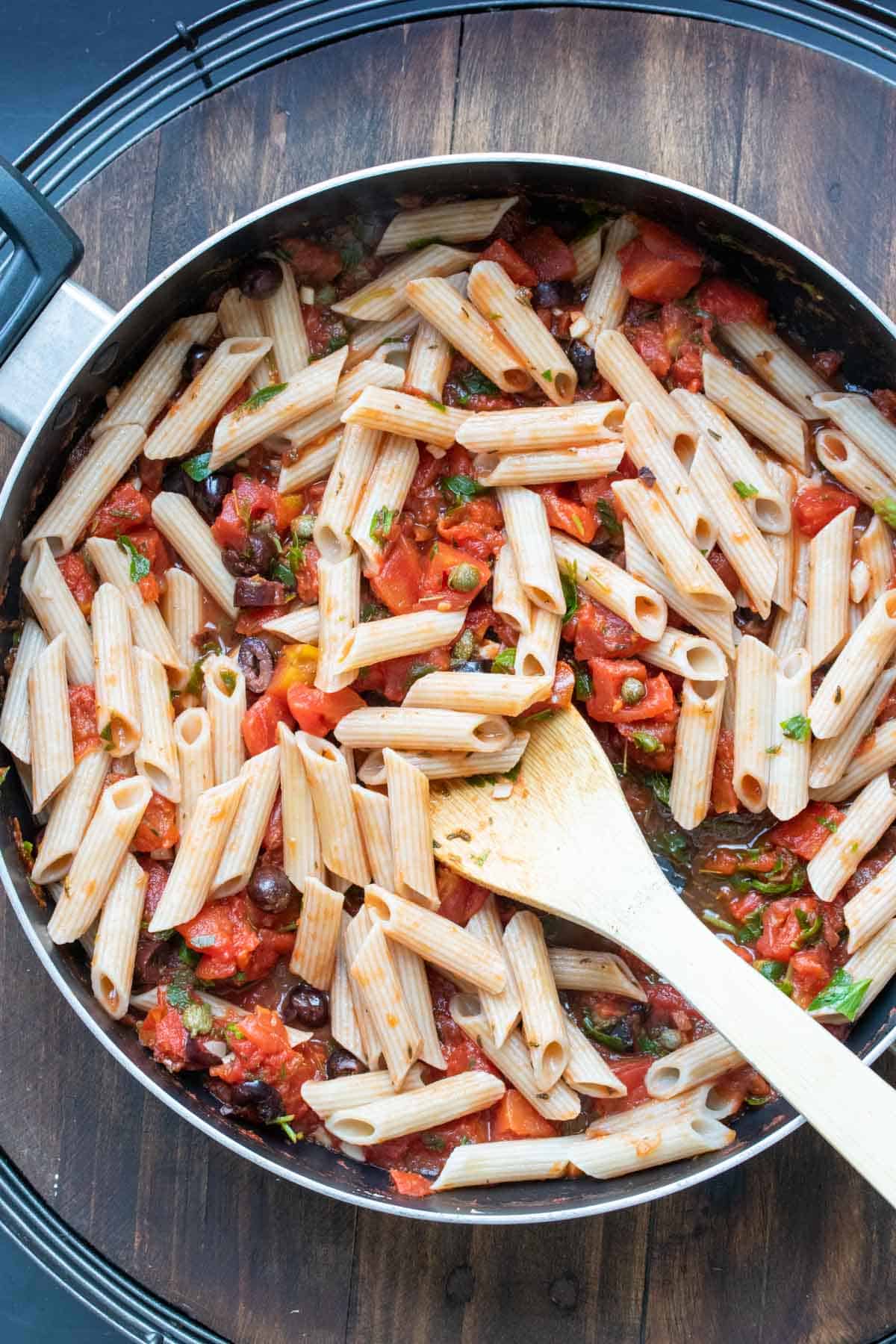 Wooden spoon mixing pasta with puttanesca sauce in a pan