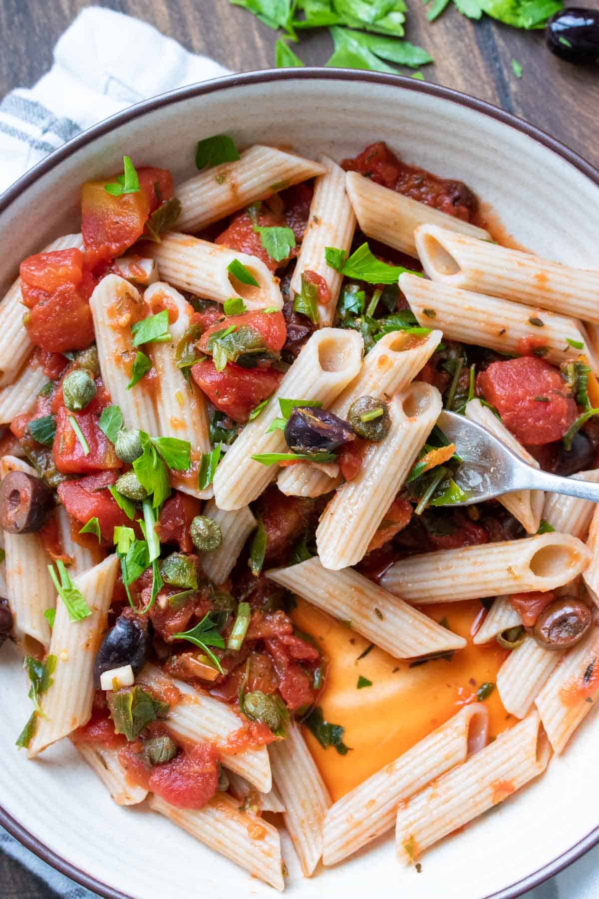 Fork taking a bite of penne pasta with puttanesca sauce from a bowl.