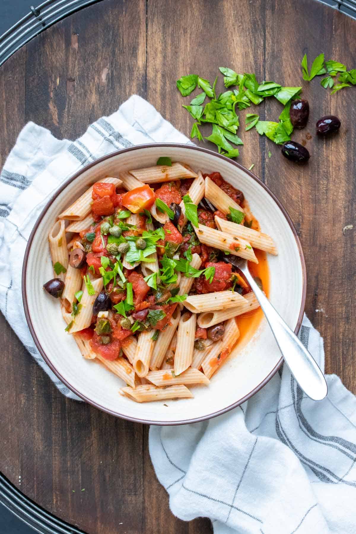 White bowl with penne pasta mixed with puttanesca sauce and parsley.