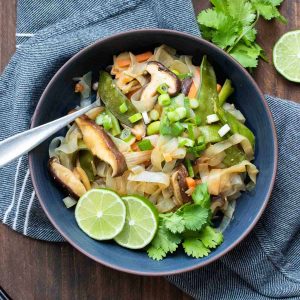 Stir fried noodles and veggies in a dark blue bowl with lime slices and cilantro