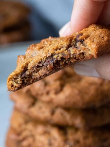 Hand holding a chocolate chip cookie with a bite out of it over a stack of cookies