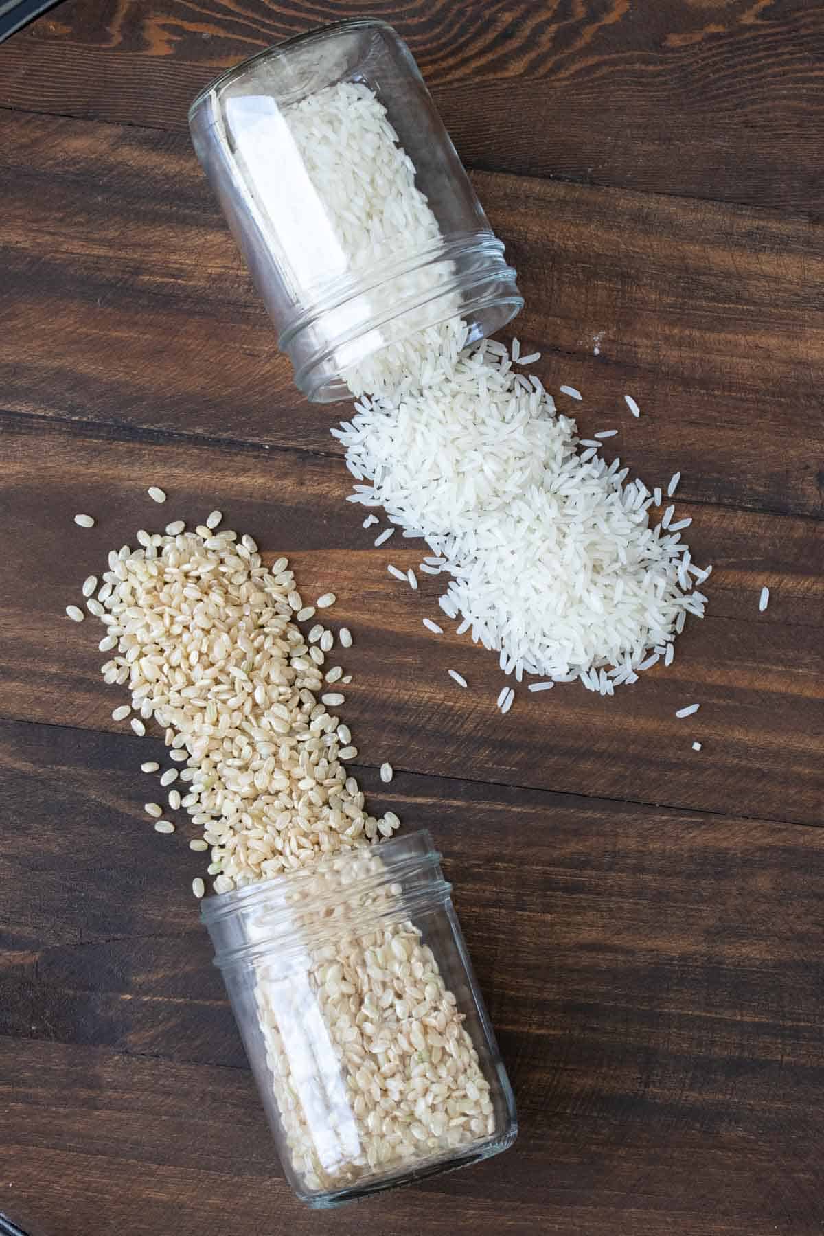 Brown and white rice spilling out of jars onto a wooden table