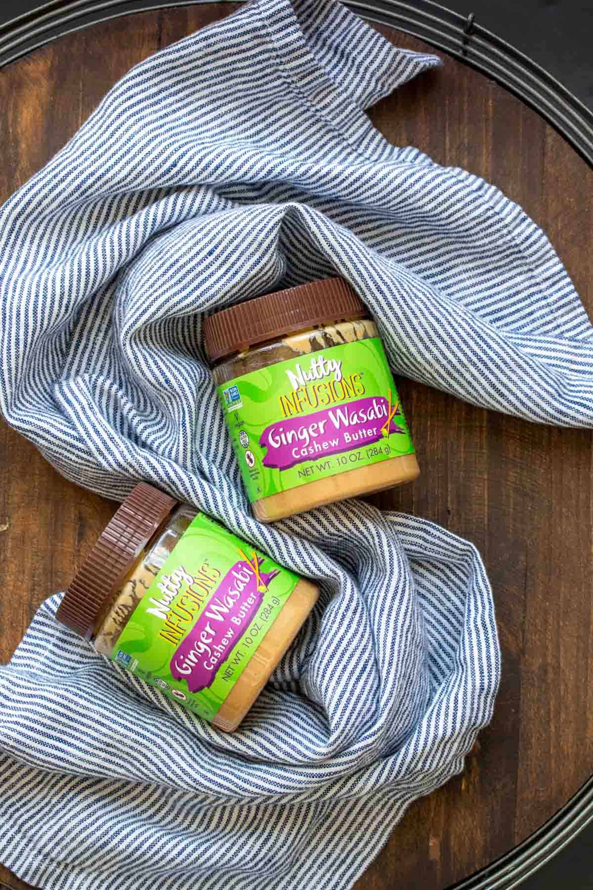 Two jars of ginger wasabi cashew butter laying next to a blue striped towel