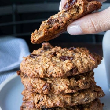 Hand holding a half eaten oatmeal raisin cookie over a stack of more