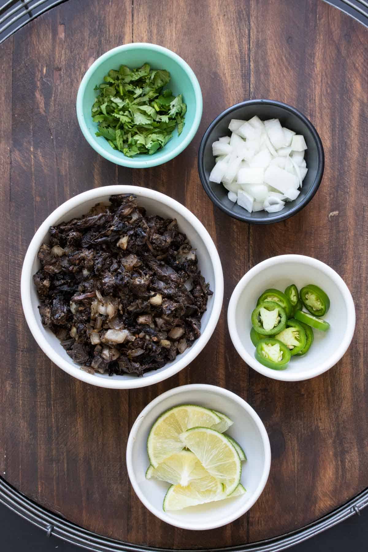 Five bowls filled with carnitas taco ingredients on a wooden table