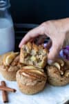 A stack of apple muffins on a plate with a hand grabbing one with a bite out of it.