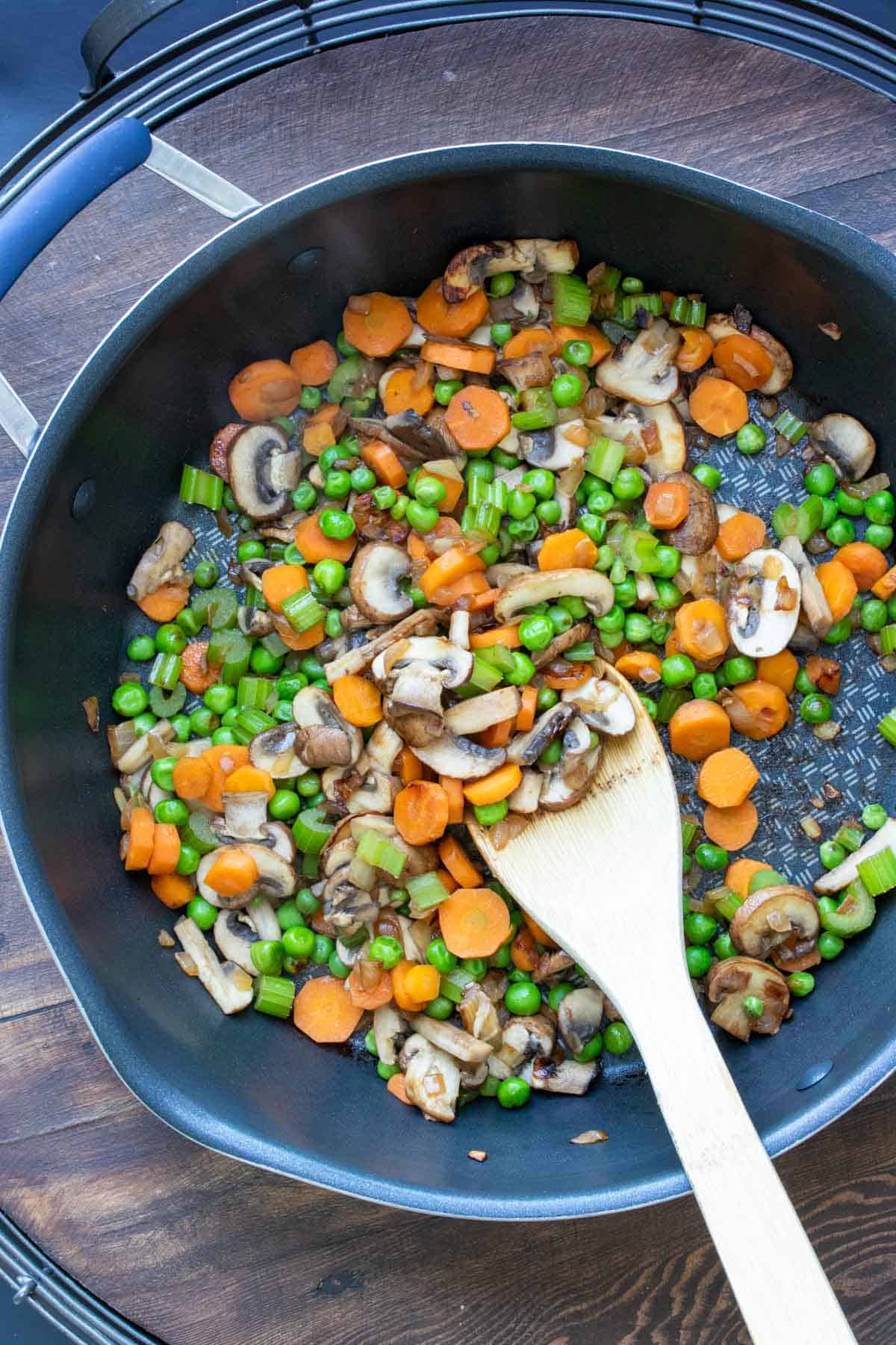 Veggies being sautéed in a pan by a wooden spoon.