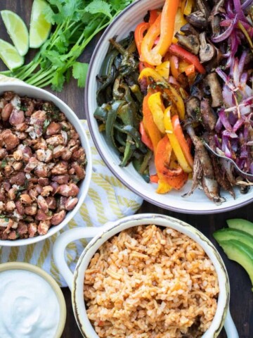 Bowls of sautéed peppers, rice and beans next to sour cream and avocado on a wooden surface