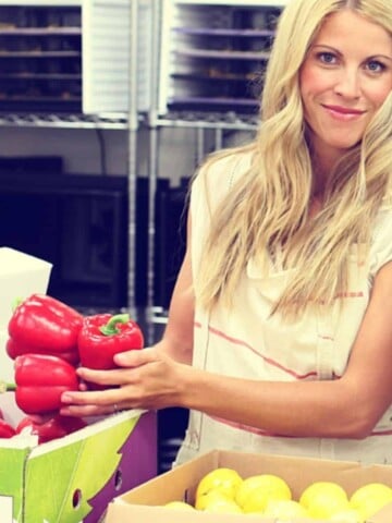 Photo of a blond woman in front of boxes of produce holding red peppers