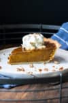 Pumpkin pie piece on a white plate topped with whipped cream and sprinkled with nuts.