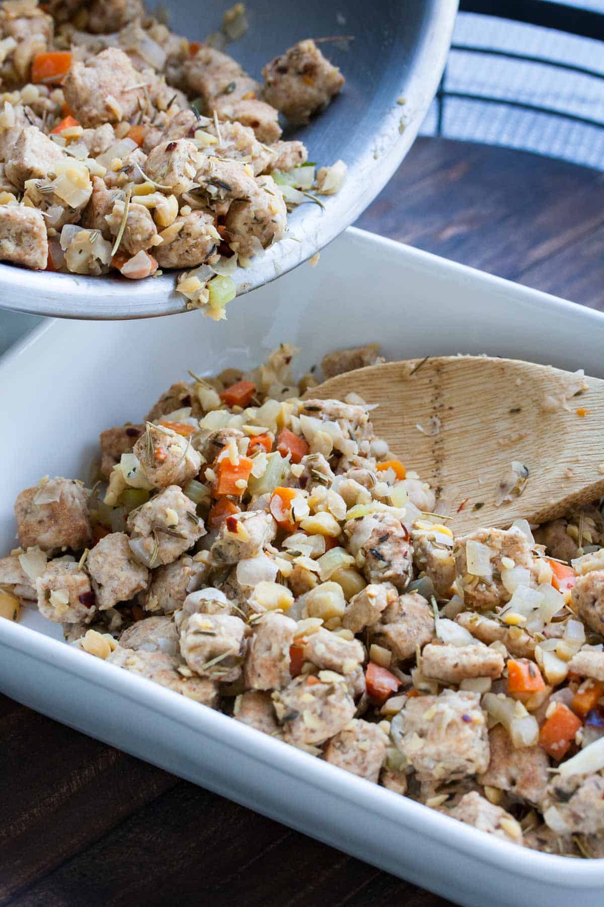 Pouring stuffing from silver bowl into white baking dish with a wooden spoon