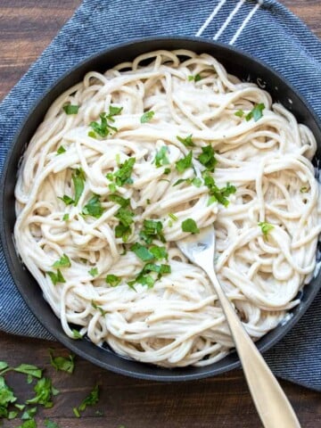 A black skillet filled with pasta noodles covered in Alfredo sauce with a fork in the middle