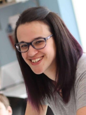 A woman with brown hair with purple ends and a grey shirt leaning over smiling