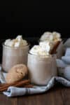 A photo of three glass jars of eggnog topped with whipped cream and a cookie next to them.