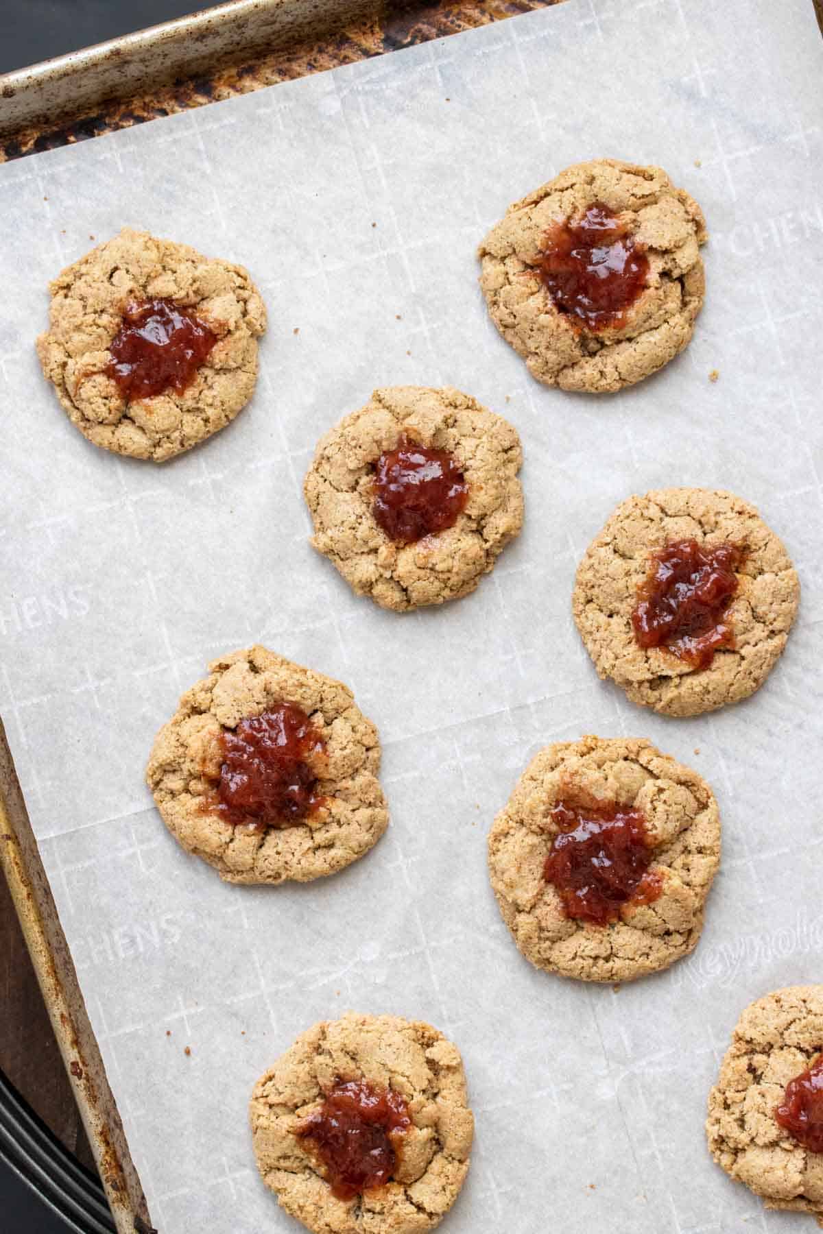 Baked thumbprint cookies with jam on a piece of parchment on a cookie sheet