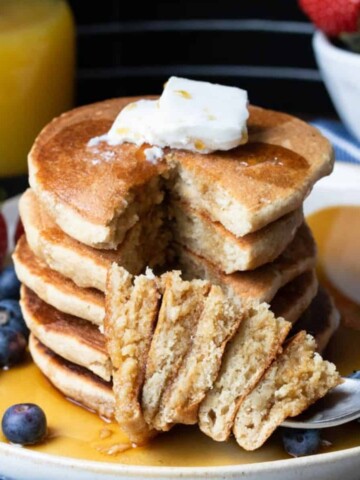 Fork with a stacked bite of pancakes in front of a big stack with syrup and butter