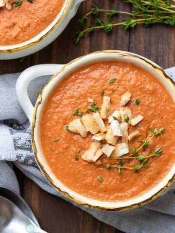 A bowl of tomato soup topped with croutons and fresh thyme springs on a wooden surface