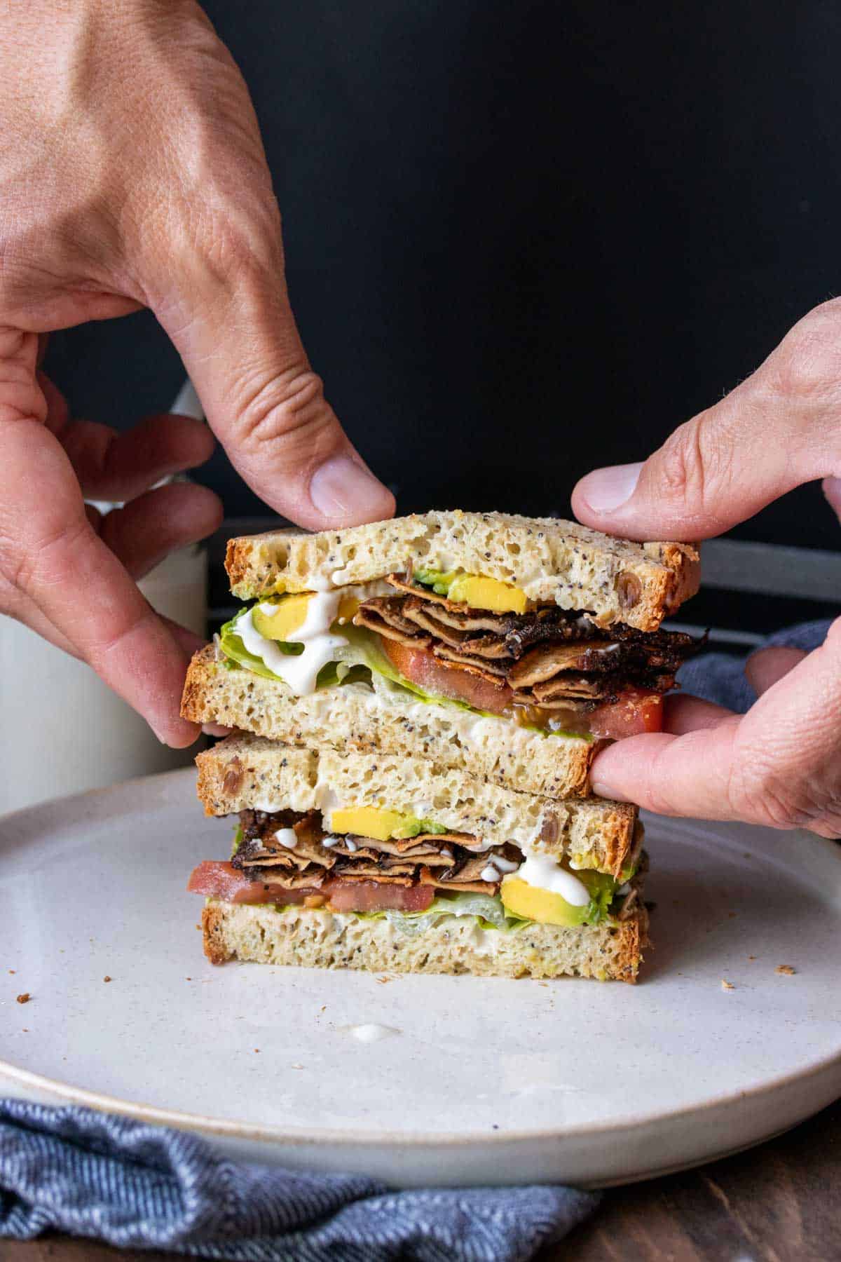 Hands grabbing half of a vegan BLT sandwich from a stack