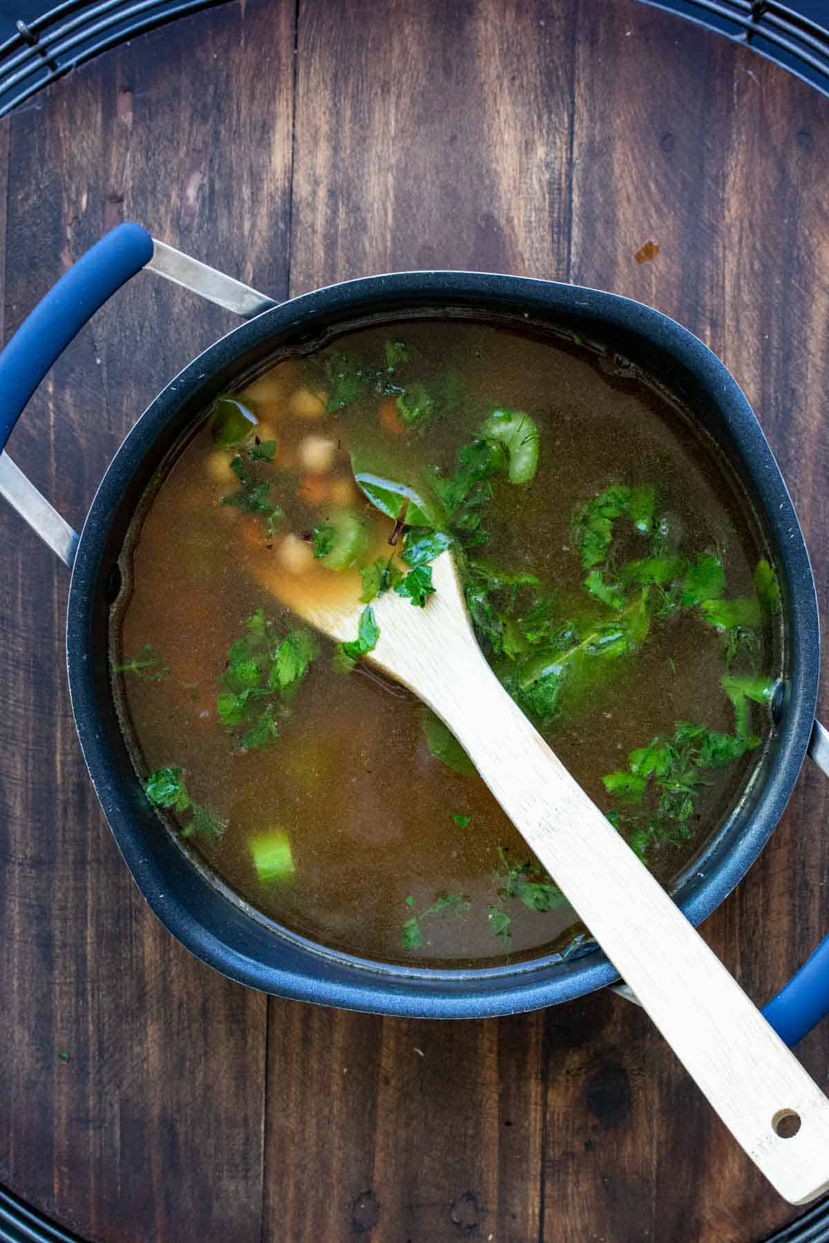 Pot with broth and veggies being stirred by a wooden spoon