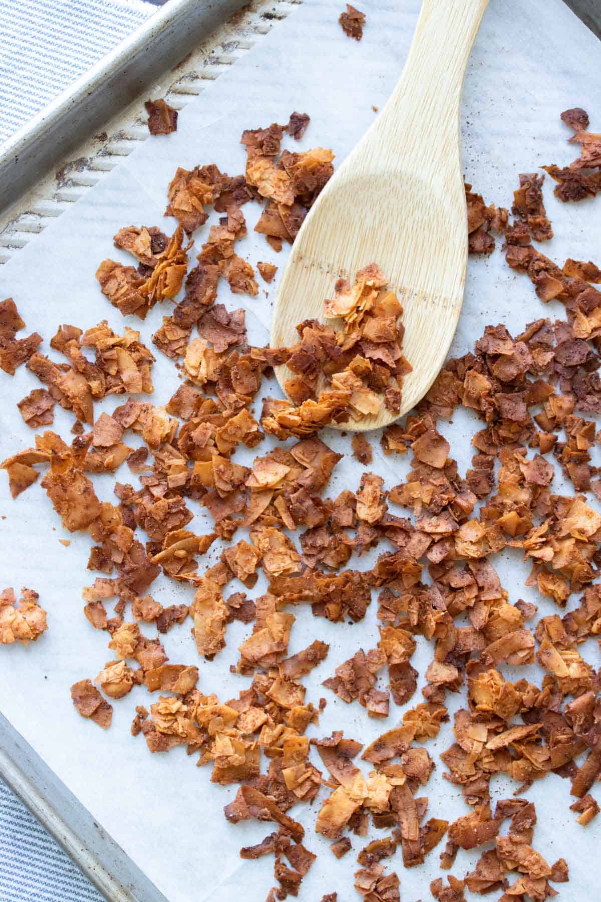 Wooden spoon stirring coconut bacon pieces on baking sheet